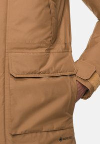 Brun Gore-Tex jakke med en stor frontlomme, glat tekstur og en tætsiddende manchet med velcro-lukning. Enkel design og funktionelle detaljer.