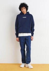 Marineblå hettegenser med hvit tekstlogo, lagd over en hvit skjorte, kombinert med mørkeblå jeans og hvite joggesko.