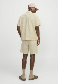 Kurzärmliges gemustertes Hemd und Shorts in hellbeige mit geometrischen Designs. Ergänzt durch eine pinke Mütze und braune Sandalen.