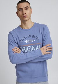 Felpa azzurra con scollatura rotonda. Presenta del testo stampato in vari colori in modo audace. Tessuto morbido e maniche lunghe con polsini a coste.