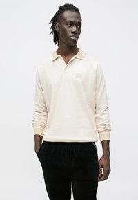 Šviesios kremo spalvos ilgomis rankovėmis polo marškinėliai su tekstūruotu raštu, smėlio spalvos apykakle ir išsiuvinėtu "OPEN ERA" logotipu kairiajame krūtinės aukštyje. Derinami su juodomis korduro kelnėmis.