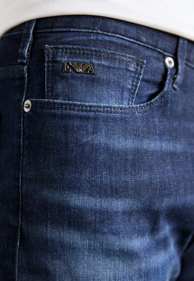 Dunkelblaue Denim-Jeans mit einer Vordertasche, die mit einem kleinen schwarzen Logo-Patch versehen ist. Der Stoff zeigt eine dezente Struktur und ein leichtes Verblassen.