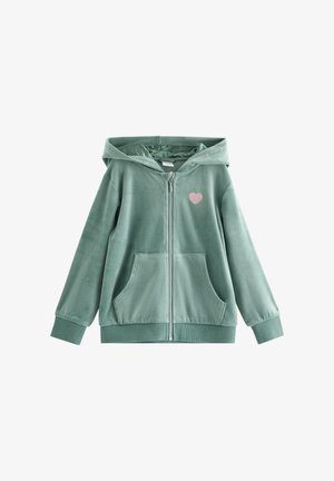 Blond pige smilende, iført grøn velour hættetrøje med lynlås og matchende bukser, blomstret skjorte og hvide sneakers, stående mod en ensfarvet baggrund.
