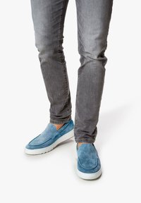 Blauwe suède instapschoenen met witte rubberen zolen, gecombineerd met grijze slim-fit jeans. De schoenen hebben een ronde neus en een gladde textuur.