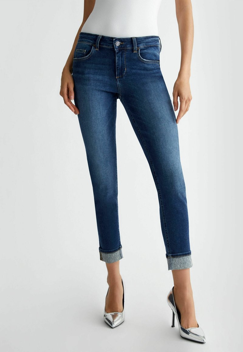 Femme portant un jean skinny bleu foncé retroussé et des chaussures à talons hauts argentées, debout devant un fond clair uni.