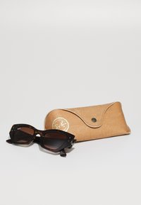 Ray-Ban napszemüveg sötét teknősbéka mintájú kerettel és barna lencsékkel, egy világosbarna bőrtok mellett, amelyen arany logó található.