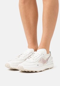 Piernas que llevan zapatillas Nike blancas y beige con un logotipo de Swoosh rosa, de pie sobre una superficie blanca con un fondo liso.