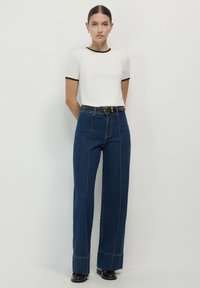 Top bianco a maniche corte con bordi neri, abbinato a jeans a gamba larga blu scuro a vita alta. Cintura nera con dettagli in oro.