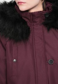 Parka maro cu glugă din blană sintetică neagră. Are închidere cu fermoar, accente cu nasturi și material texturat. Include un buzunar cu petic pe lateral.