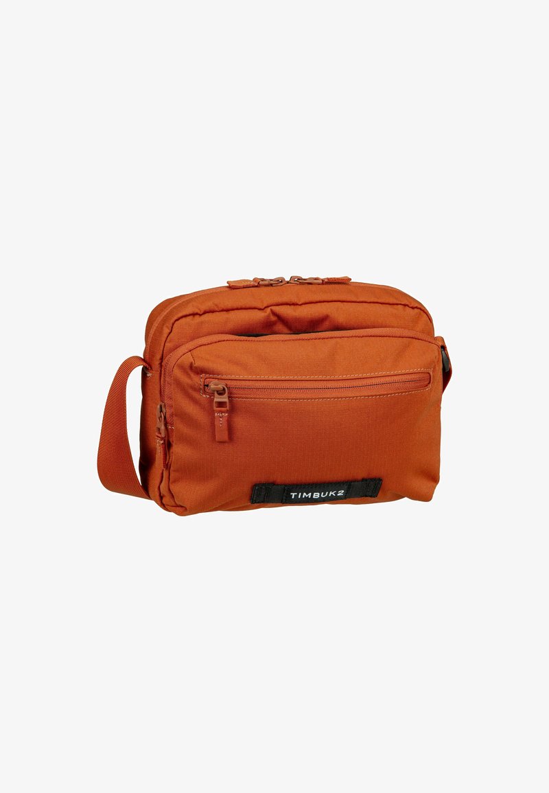 Bolso mensajero de lona naranja con un bolsillo frontal con cremallera, correa ajustable y parche con logo. Presenta una textura suave y un diseño minimalista.