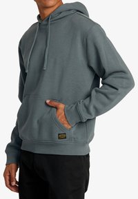 Sudadera con capucha en un tono azul verdoso suave, hecha de tela blanda. Cuenta con un bolsillo canguro en la parte frontal, puños acanalados y un parche con el logotipo cerca del dobladillo.