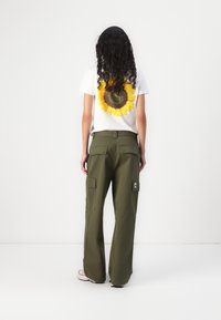 Weißes T-Shirt mit einem Sonnenblumen-Graphic auf der Rückseite, kombiniert mit olivgrünen Cargo-Hosen, die mehrere Taschen und ein aufgenähtes Logo haben.