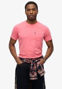 Camiseta de algodón rosa con un bolsillo en el pecho, combinada con jeans oscuros y una camisa de cuadros atada a la cintura. Diseño simple y ajustado.