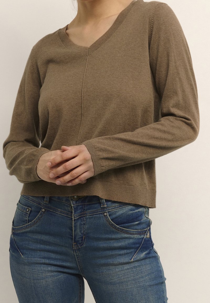 Pull en maille marron avec un col en V, des manches longues et des poignets côtelés. Associé à un jean en denim bleu, mettant en avant un look décontracté.