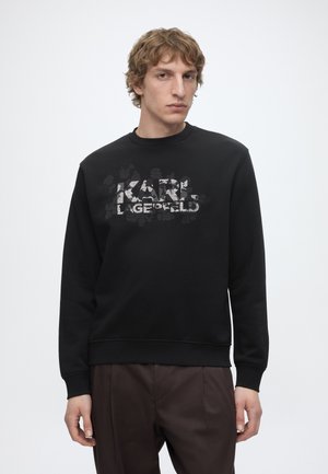 Młody mężczyzna z kręconymi włosami ubrany w czarną bluzę z teksturowanym logo "KARL LAGERFELD", połączoną z ciemnobrązowymi spodniami.