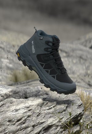 Botte de randonnée imperméable Columbia avec semelle Vibram suspendue au-dessus d'un terrain rocheux, mettant en avant un design robuste de chaussure d'extérieur.