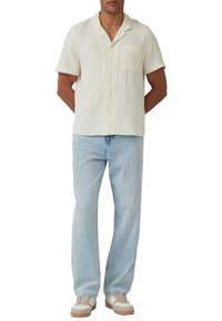 Crème kleurig overhemd met korte mouwen, knoopsluiting en een gestructureerde stof, gecombineerd met lichtblauwe rechte jeans en witte sneakers.