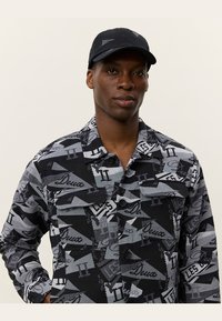 Cappello nero e camicia a fantasia con forme geometriche grigie e nere, elementi di testo e due tasche sul petto, realizzata in un tessuto con finitura texture.