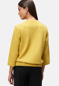 Haut jaune texturé avec des manches 3/4, un col rond et un ourlet froncé. Porté sur un pantalon noir, montrant un subtil motif de tissage.
