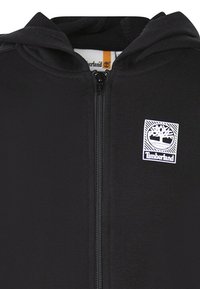 Felpa nera con zip realizzata in tessuto liscio, dotata di zip frontale, cappuccio attaccato e patch logo Timberland sul lato sinistro del petto.