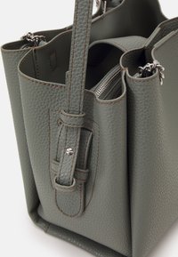 Šedá texturovaná kožená taška přes rameno s dvěma uchy, boční zipová kapsa a kontrastní hnědé švy podél okrajů.