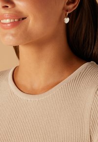 Boucle d'oreille en or avec un accent en forme de cœur blanc. Portée avec un haut texturé beige à côtes présentant un col rond.