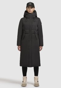 Crni dugi zimski kaput s visokim ovratnikom, prednjim džepovima i odvojivom kapuljačom, nosi se s crnom kapom i krem-bijelim tenisicama.