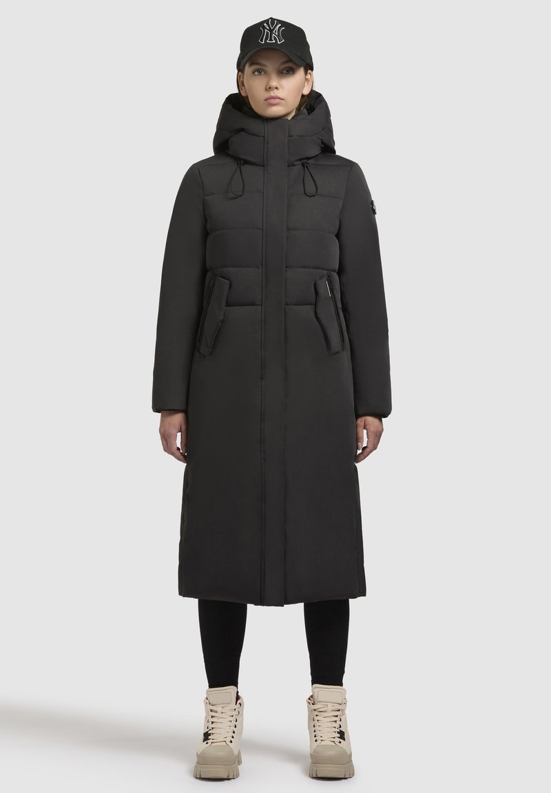 Crni dugi zimski kaput s visokim ovratnikom, prednjim džepovima i odvojivom kapuljačom, nosi se s crnom kapom i krem-bijelim tenisicama.