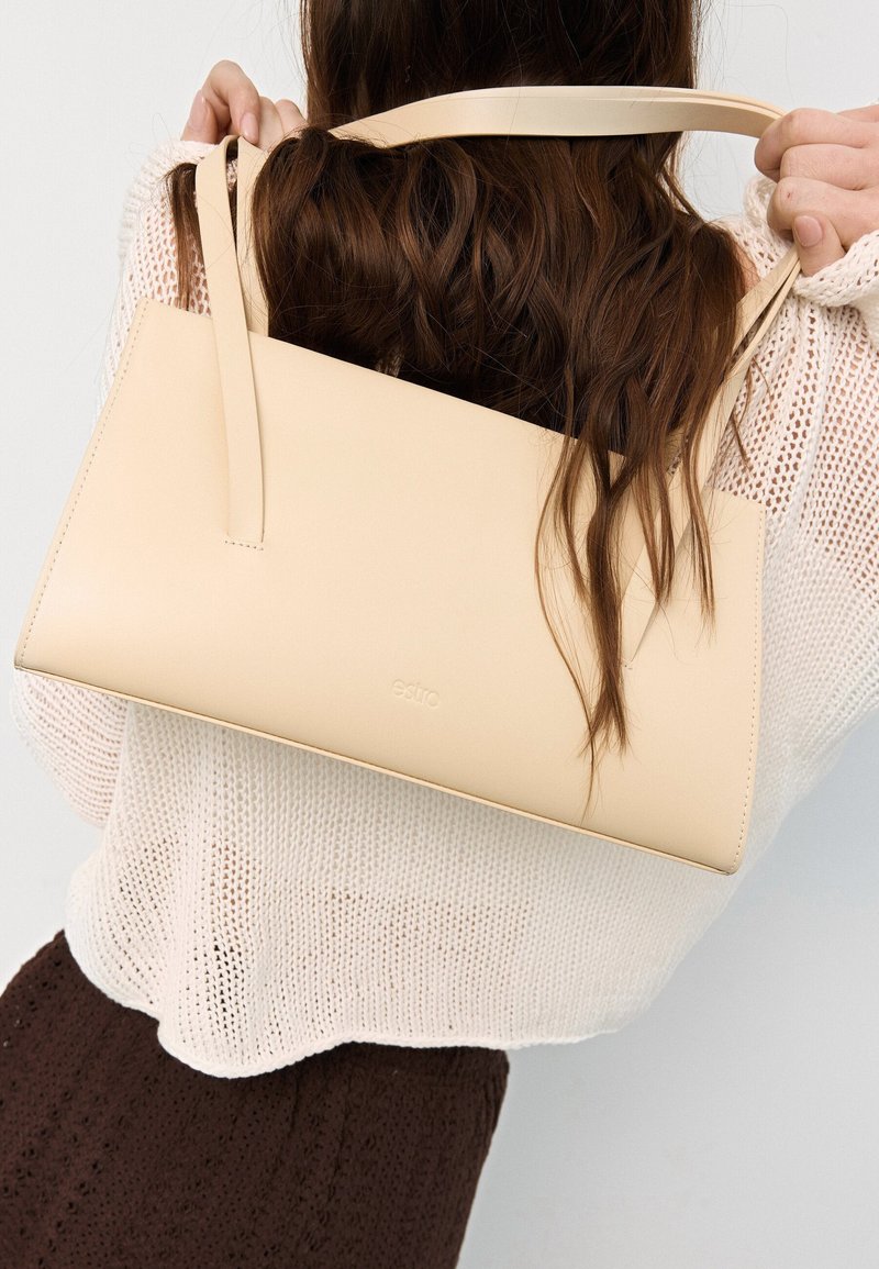 Beige Lederhandtasche mit rechteckiger Form, flachem Design, schmalen Trägern und dezent geprägtem Branding, gehalten von einer Person.