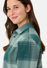 Karierter Button-Up-Hemd in verschiedenen Blau- und Grüntönen, mit Kragen, sichtbaren Knöpfen und einem gestickten Logo auf dem Ärmel.