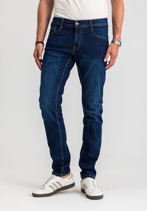 Ciemnoniebieskie dżinsy o dopasowanym kroju, z pomarańczowym przeszyciem, przednimi kieszeniami i średnim stanem. Noszone z białymi butami sportowymi.