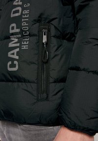Chaqueta acolchada negra con una superficie texturizada, que cuenta con una cremallera frontal y un logotipo impreso. Incluye puños elásticos y un dobladillo con cordón.