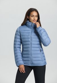 Femme portant une veste légère bleu clair matelassée avec un col montant, zippée, se tenant avec une main près de son cou et l'autre à ses côtés.