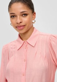 Helles pinkes Blusenshirt mit Kragen, Knopfleiste und vertikalen Falten an den Schultern. Weicher, strukturiertem Stoff mit lockerer Passform.