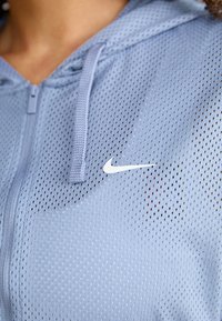 Chaqueta de malla azul claro con cremallera y capucha, que presenta el logo de la marca Nike en blanco en el pecho y una perforación texturizada en toda la tela.