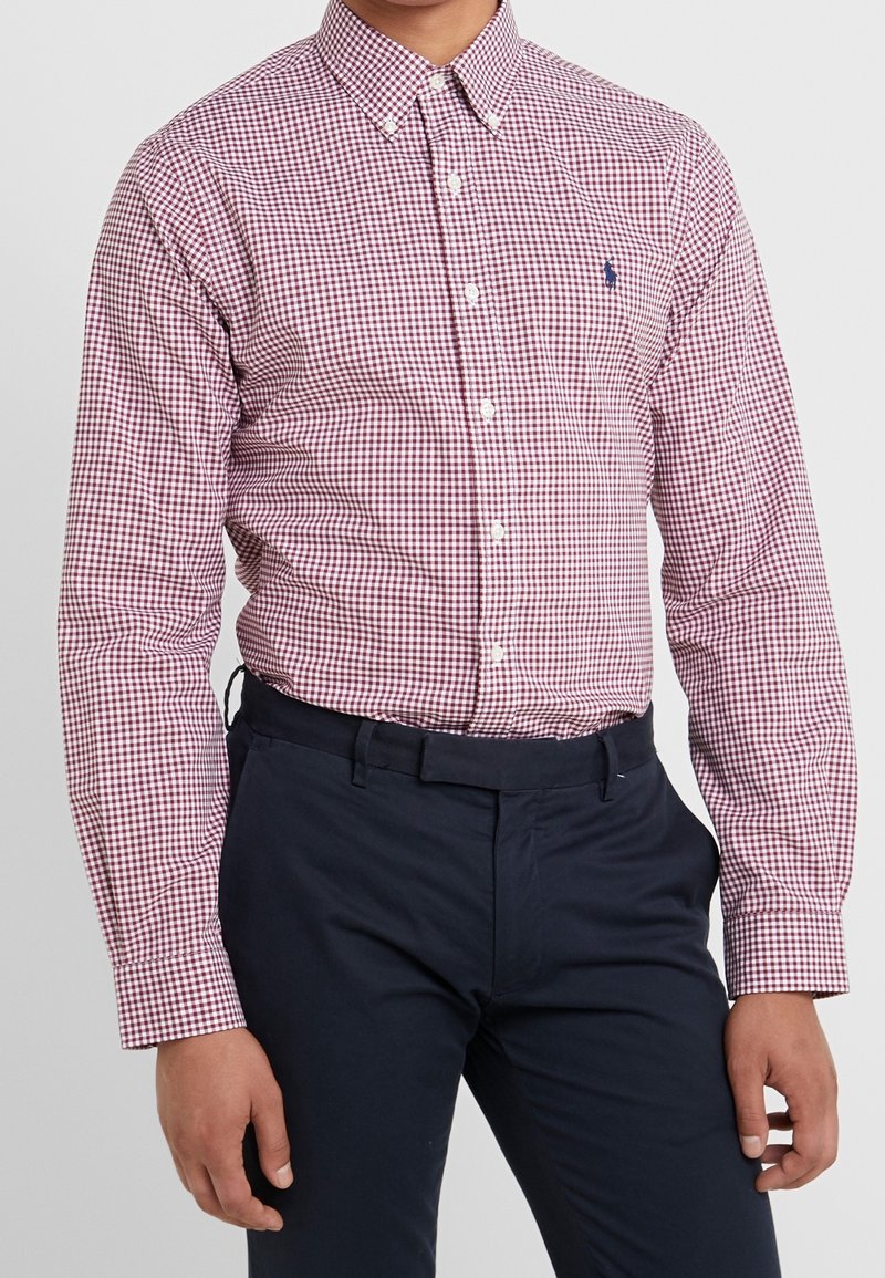 Man i en röd och vit rutmönstrad skjorta med knappar, instoppad i mörkblå byxor, som står med avslappnade händer vid sidorna.