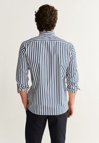 Camicia a righe con righe verticali blu navy e bianche, maniche lunghe con polsini arrotolati, colletto con bottoni e orlo curvo.