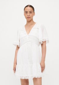 Abito corto bianco con scollatura a V, maniche a sbuffo, dettagli in pizzo e orlo arricciato. Presenta chiusure con bottoni e tessuto testurizzato con motivi floreali.