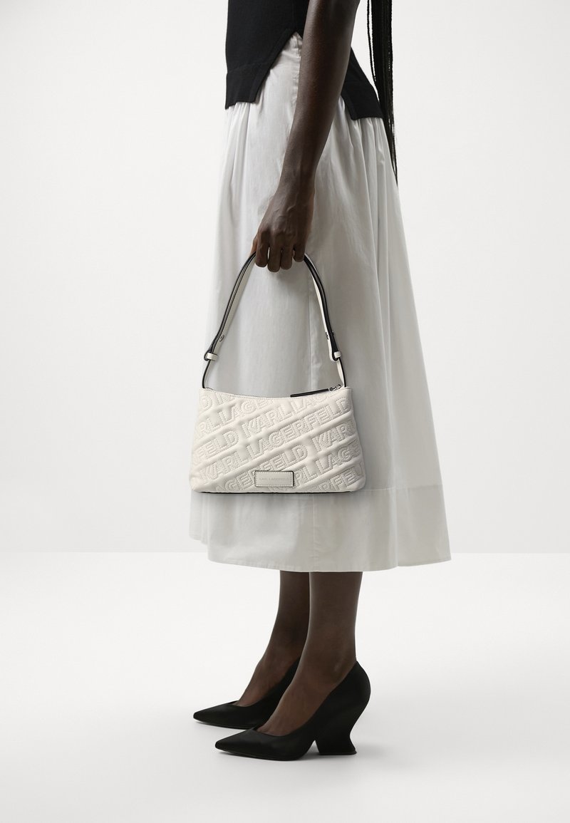 Femme portant une jupe midi blanche et des talons noirs, tenant un petit sac à main blanc embossé par sa poignée à son côté.