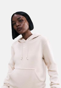 Felpa beige con coulisse e tasca frontale. Presenta un piccolo logo Nike bianco sul petto, realizzata in tessuto morbido e texture.
