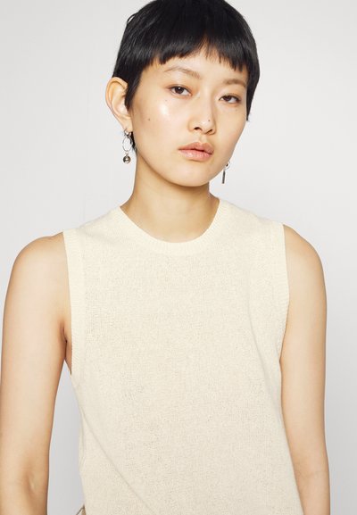 Fiatal személy rövid fekete hajjal, ujjatlan krém színű kötött felsőben és aszimmetrikus ezüst fülbevalókkal, semleges arckifejezéssel, sima háttér előtt.