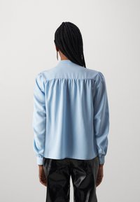 Lyseblå bluse med rynket bak, lange ermer og en myk, glatt tekstur. Kombinert med blanke svarte bukser.
