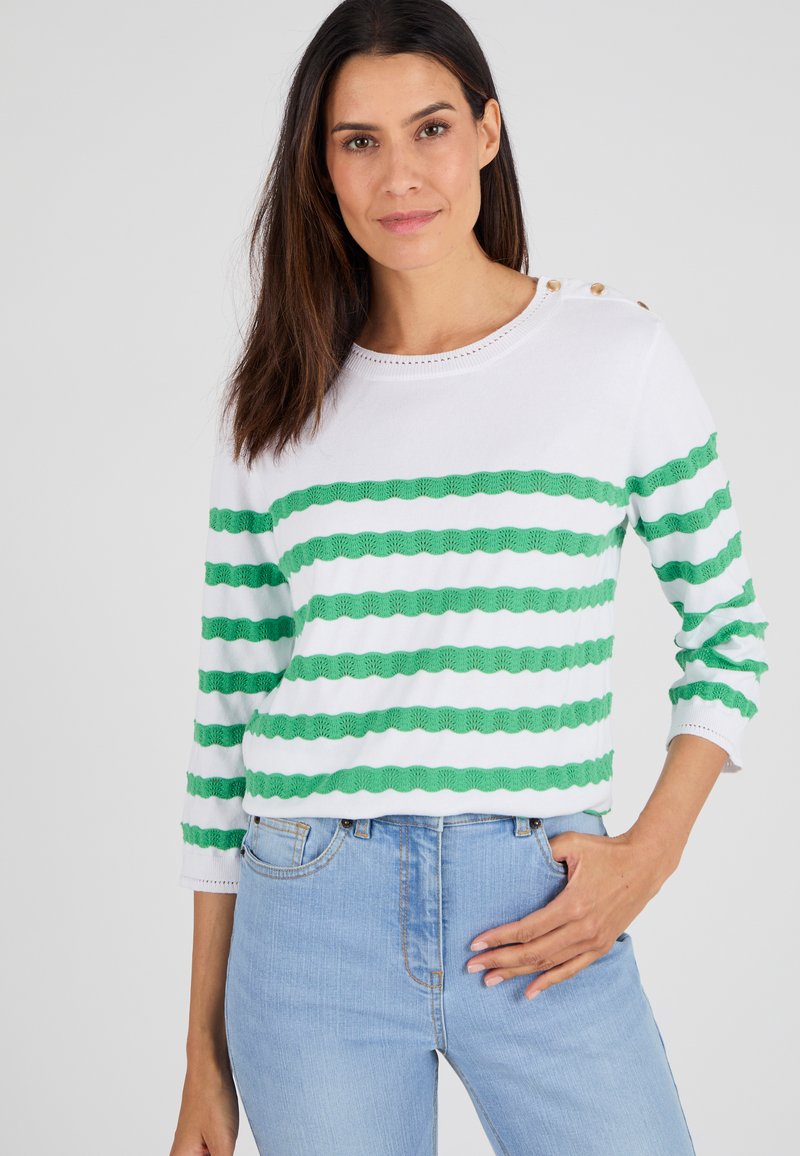 Witte gebreide trui met groene horizontale strepen en golfpatronen, ronde halslijn en decoratieve knopen op de schouder. Gecombineerd met blauwe jeans.