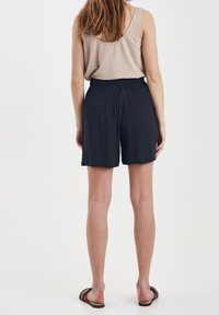 Kvinna stående med ryggen mot kameran, klädd i beige ärmlös topp, svarta shorts och bruna sandaler mot en enfärgad vit bakgrund.