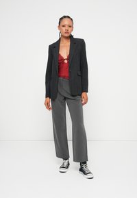 Blazer noir à rayures sur un top en dentelle rouge, associé à un pantalon large gris. Le mannequin porte des baskets noir et blanc. Fond blanc uni.