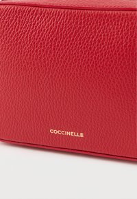 Piros bőr táska texturált felülettel. Az egyik oldalán aranyozott, dombornyomott "COCCINELLE" logóval, téglalap alakú, minimalista dizájnnal.