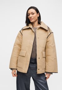 Μπεζ καπιτονέ μπουφάν με μεγάλο γιακά, μαύρο περιθώριο και δύο μπροστινές τσέπες, φορεμένο πάνω από υφασμάτινο καφέ πουλόβερ και σκούρο παντελόνι.