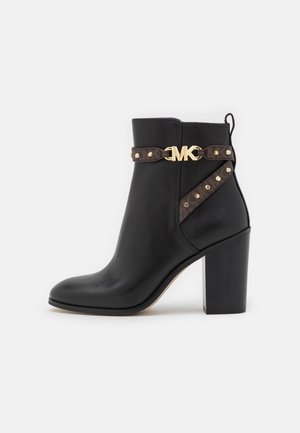 Bottine en cuir noir à bout carré, avec un talon chunky, un matériel en métal doré portant le logo MK, et une sangle décorative marron avec des rivets.