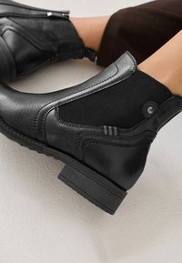 Černé kožené kotníkové boty s texturovanými tkaninovými panely, zaoblenou špičkou, nízkým blokovým podpatkem a bočním zipem. Gumová podrážka s horizontálními pruhy.