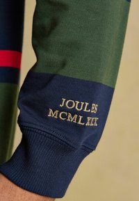 Pruhovaný zelený a námornícky modrý rukáv s výšivkou "JOULES MCMLXIX" v béžovej farbe neďaleko manžety na neformálnom odeve.